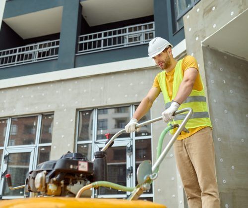 Effort, fatigue. Young adult tired man in protective helmet and bright vest with special equipment working on construction site against background of new building
